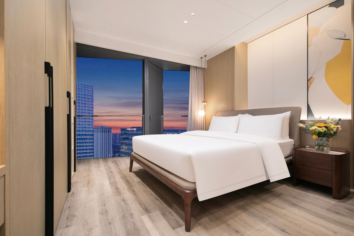 Een moderne hotelkamer met een groot bed, een nachtkastje en uitzicht op een stadsbeeld in de zonsondergang.