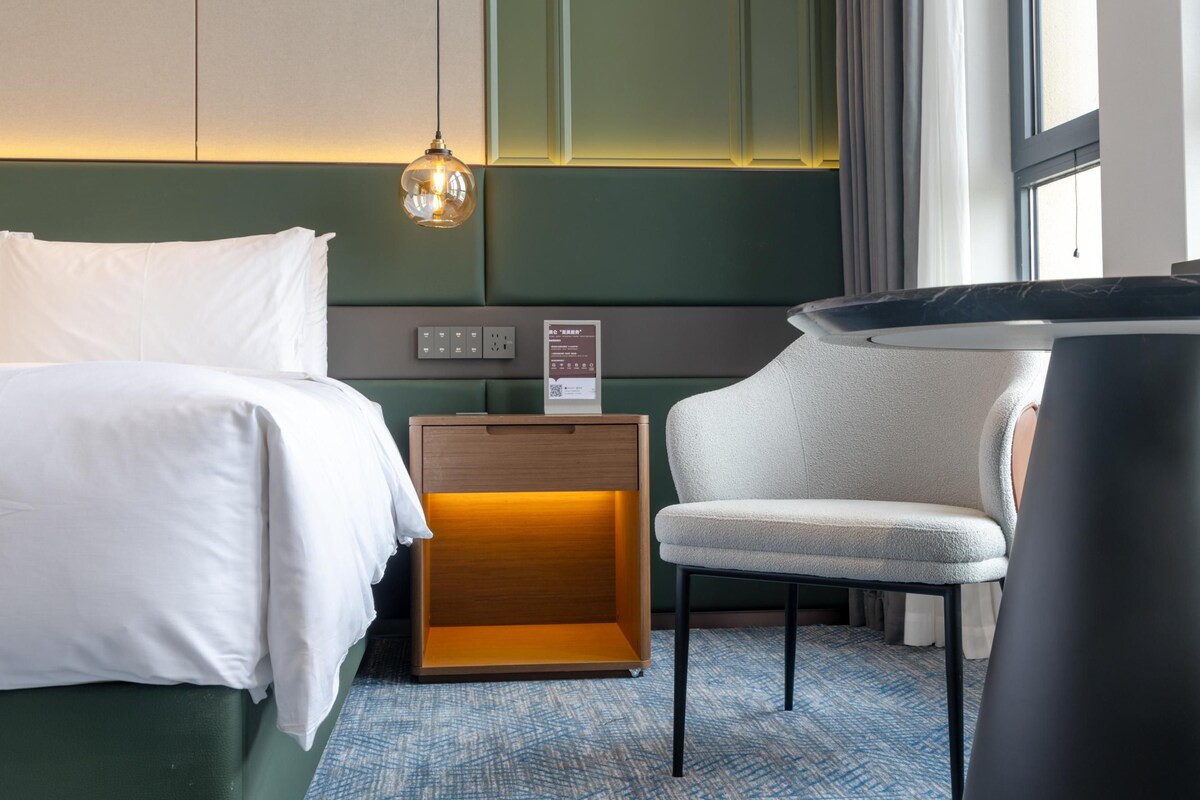 A hotel room with a bed, a chair, a nightstand, and a wall-mounted light fixture.