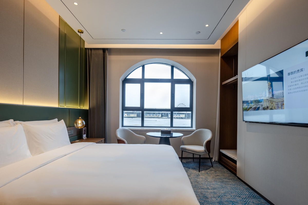 Une chambre d’hôtel avec un grand lit, un bureau avec une chaise, une grande fenêtre en arc de cercle et un écran fixé au mur qui affiche des informations.