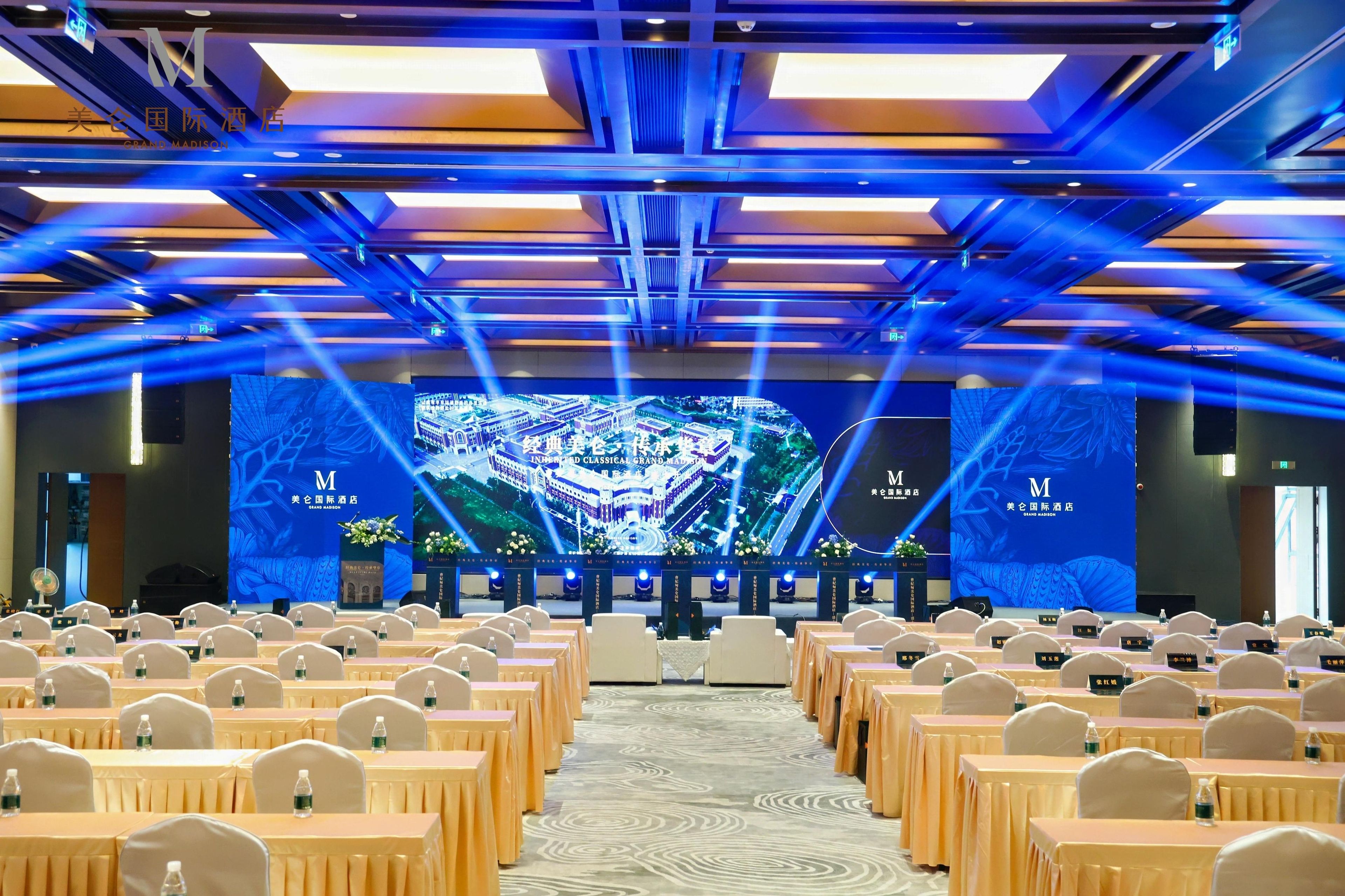 A conference room with rows of tables and chairs, a stage with a large screen displaying a building image, and a ceiling with blue lighting.