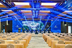 Un salón de conferencias con filas de mesas y sillas, un escenario con una gran pantalla mostrando una imagen de un edificio, e iluminación azul en el techo.