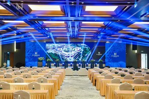 Un salón de conferencias con filas de mesas y sillas, un escenario con una gran pantalla mostrando una imagen de un edificio, e iluminación azul en el techo.