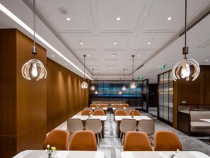 Un salón de conferencias moderno con mesas largas y sillas anaranjadas, con iluminación colgante y vistas a un paisaje urbano a través de amplios ventanales.