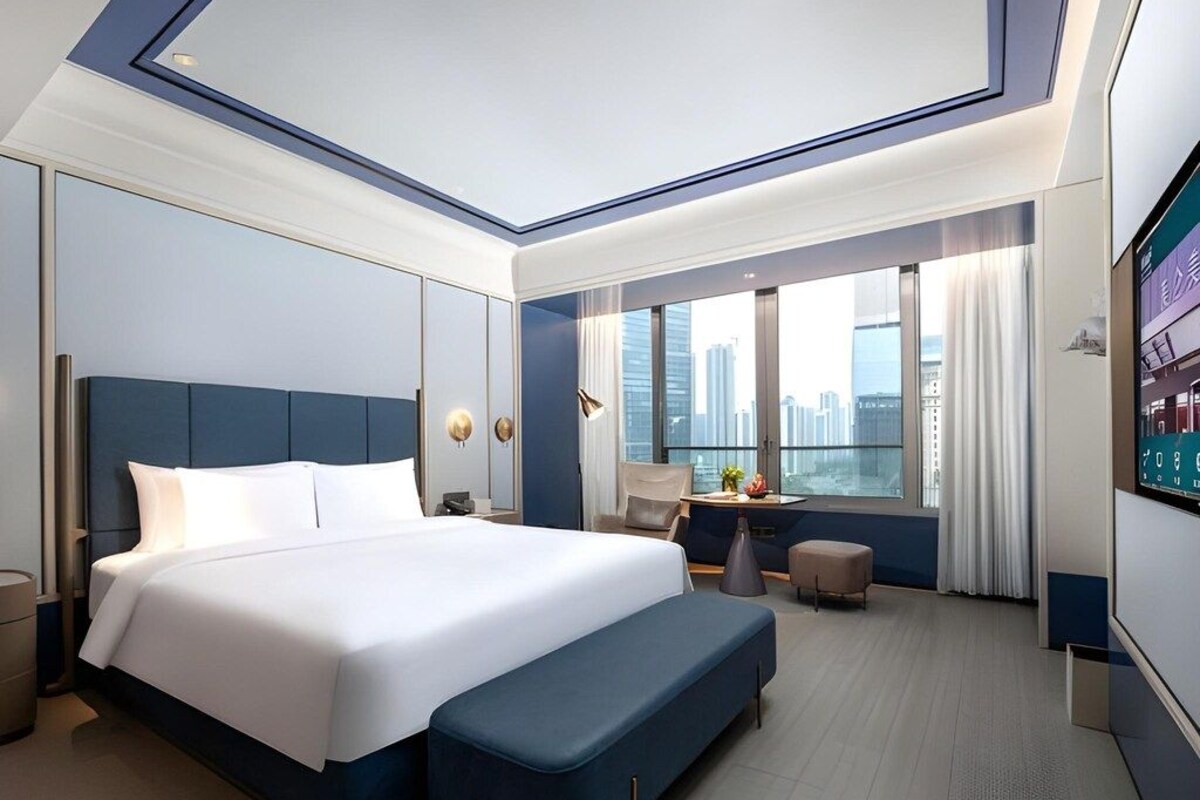 A modern hotel room with a large bed, a bench, a desk, and a city view.