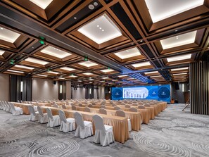 Un salón de conferencias con filas de mesas y sillas, un escenario con una pantalla grande y un diseño de techo moderno.