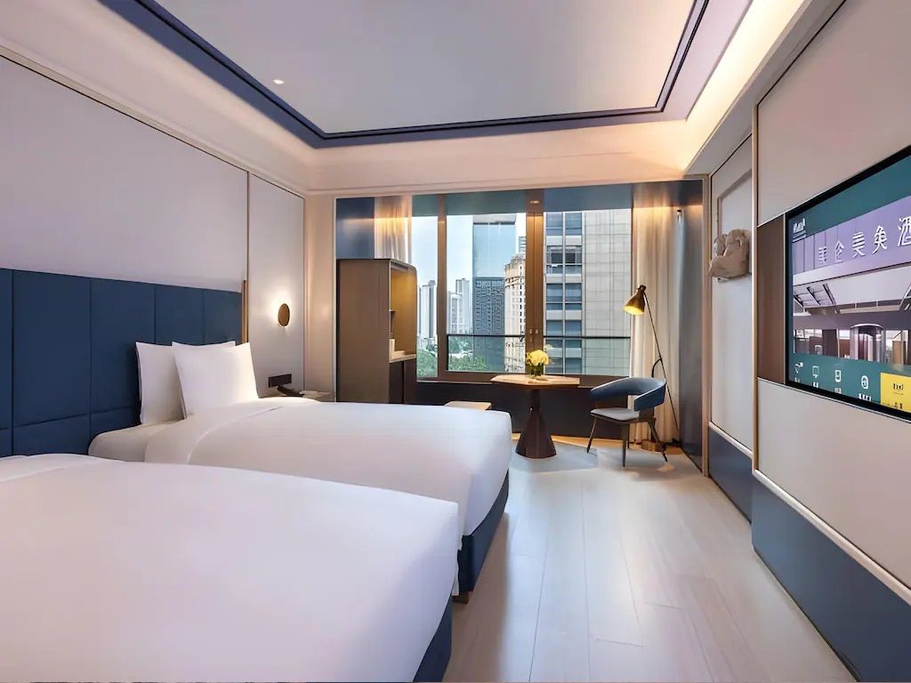 A modern hotel room with two beds, a desk, a chair, a TV, and a city view.