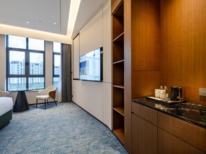 Une chambre d’hôtel avec une grande fenêtre, un lit, un bureau, une chaise, une télévision et un mini-bar.