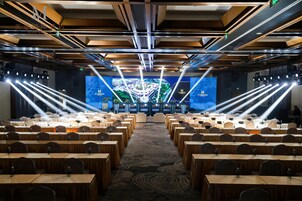 Un salón de conferencias con filas de mesas y sillas, un escenario con una pantalla grande e iluminación en el techo.