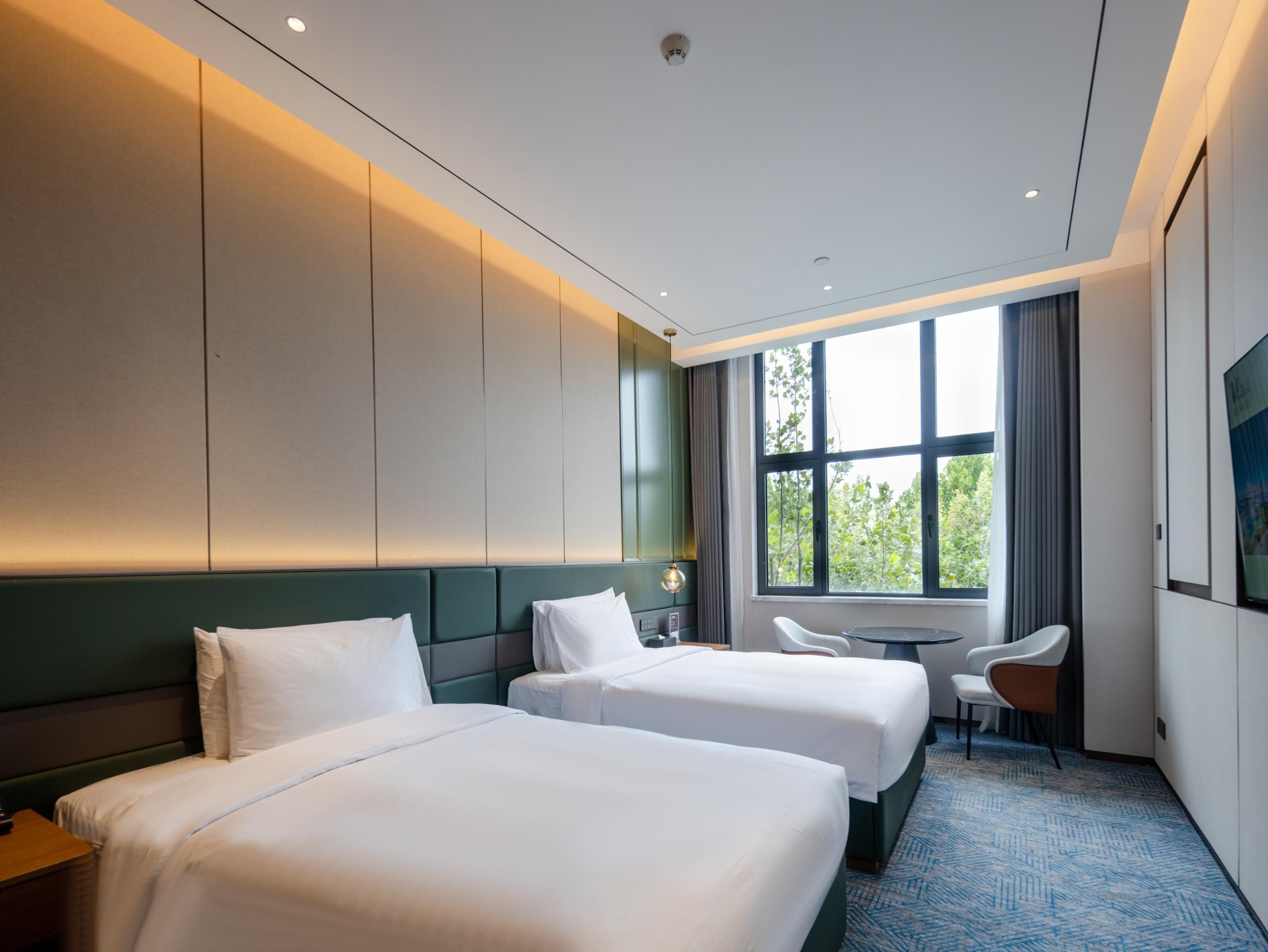 A hotel room with two beds, a desk, a chair, and a large window with a view of trees.