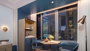 Une chambre d’hôtel moderne dotée d’une grande fenêtre, d’un bureau, de deux chaises, d’une bibliothèque et d’une lampe.