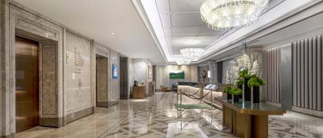 A modern hotel lobby with a geometric patterned floor, a reception desk with a glass top, and a large chandelier.