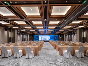 Een congreszaal met rijen tafels en stoelen, een groot scherm aan de voorkant en een kamerbreed tapijt met dessin.