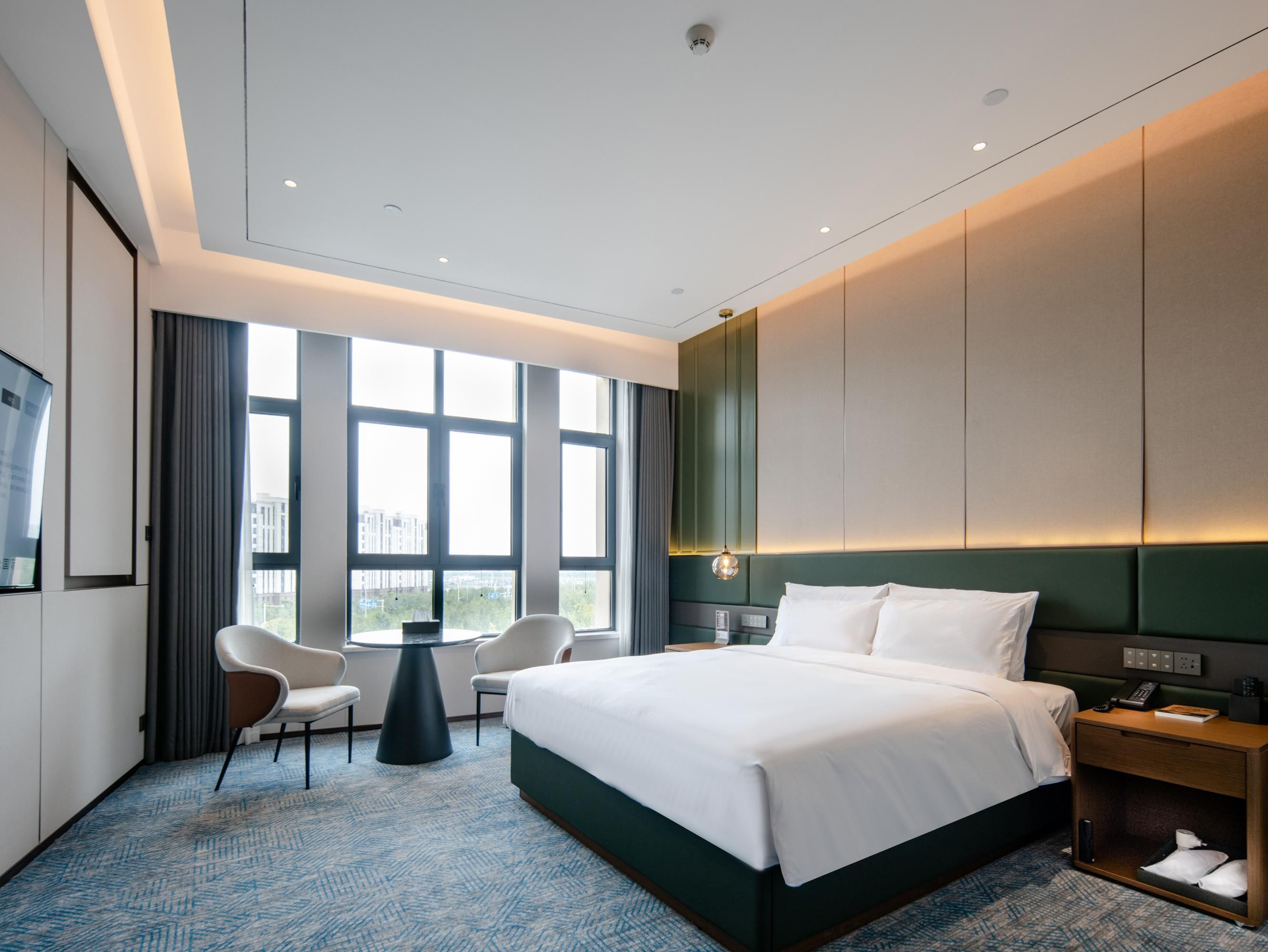 A modern hotel room with a large bed, a desk, a chair, and a view of the city through large windows.