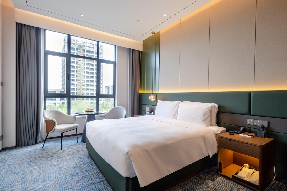 A modern hotel room with a large bed, a desk, a chair, and a view of the city through large windows.