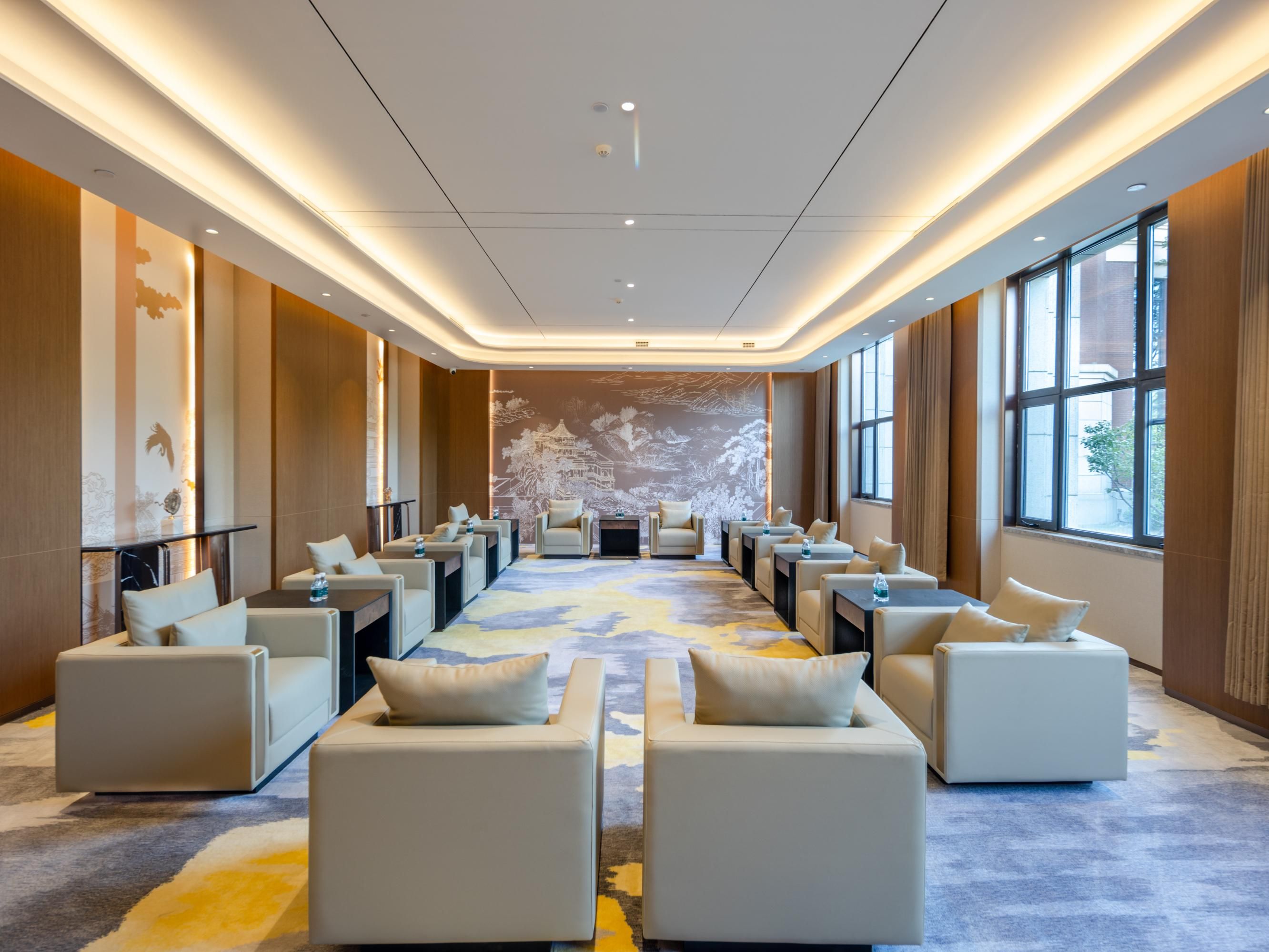 A modern hotel room with a long row of sofas and tables, large windows, and a mural on the back wall.