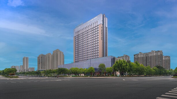 A modern high-rise building with a glass facade and a rectangular shape, surrounded by other high-rise buildings and a wide street. - Grand Madison Hotel Nantong Tongzhou High-tech (Nantong)