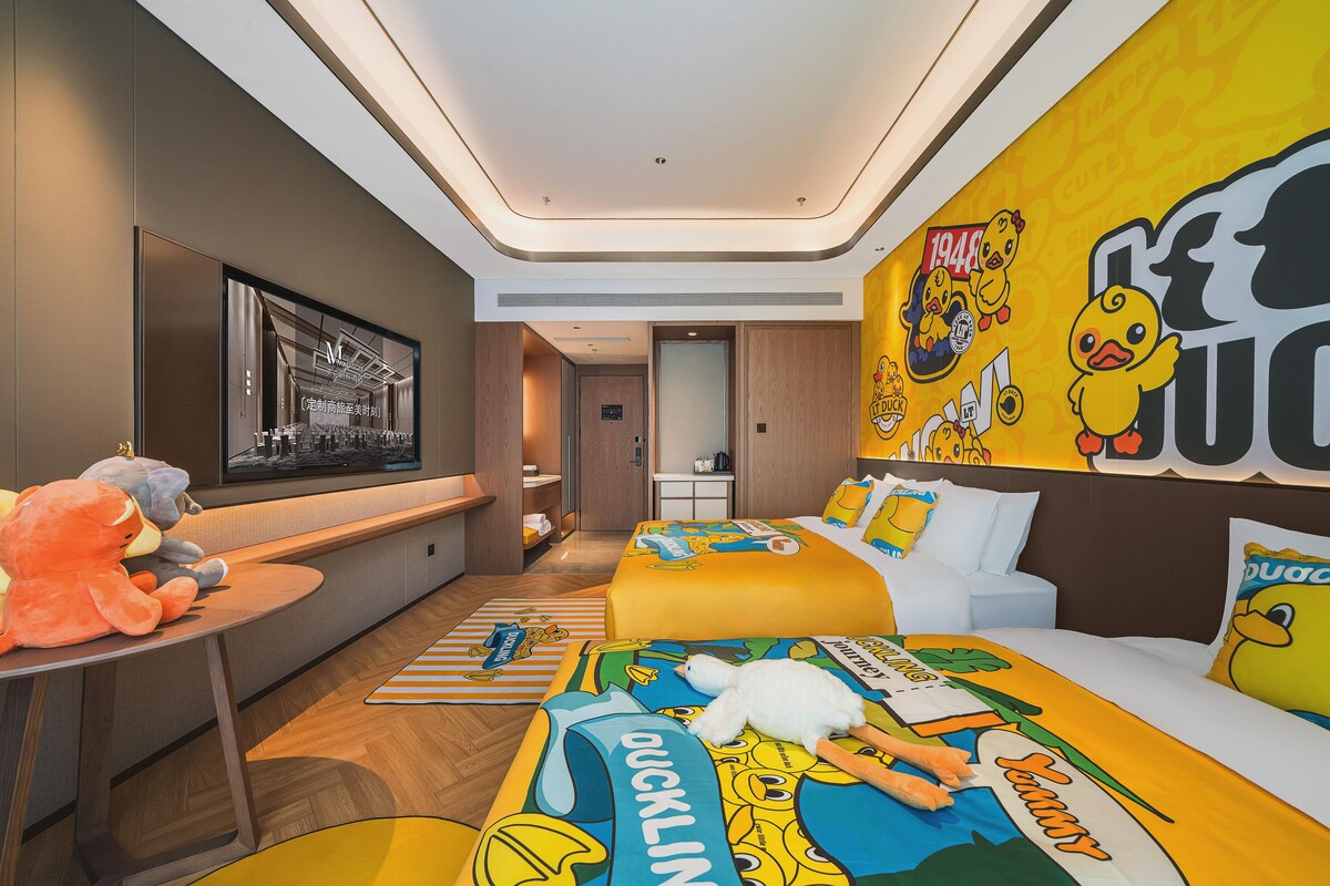 A hotel room with a modern design, featuring a large bed with duckling-themed bedding, a desk with plush toys, and a wall decorated with duckling stickers.