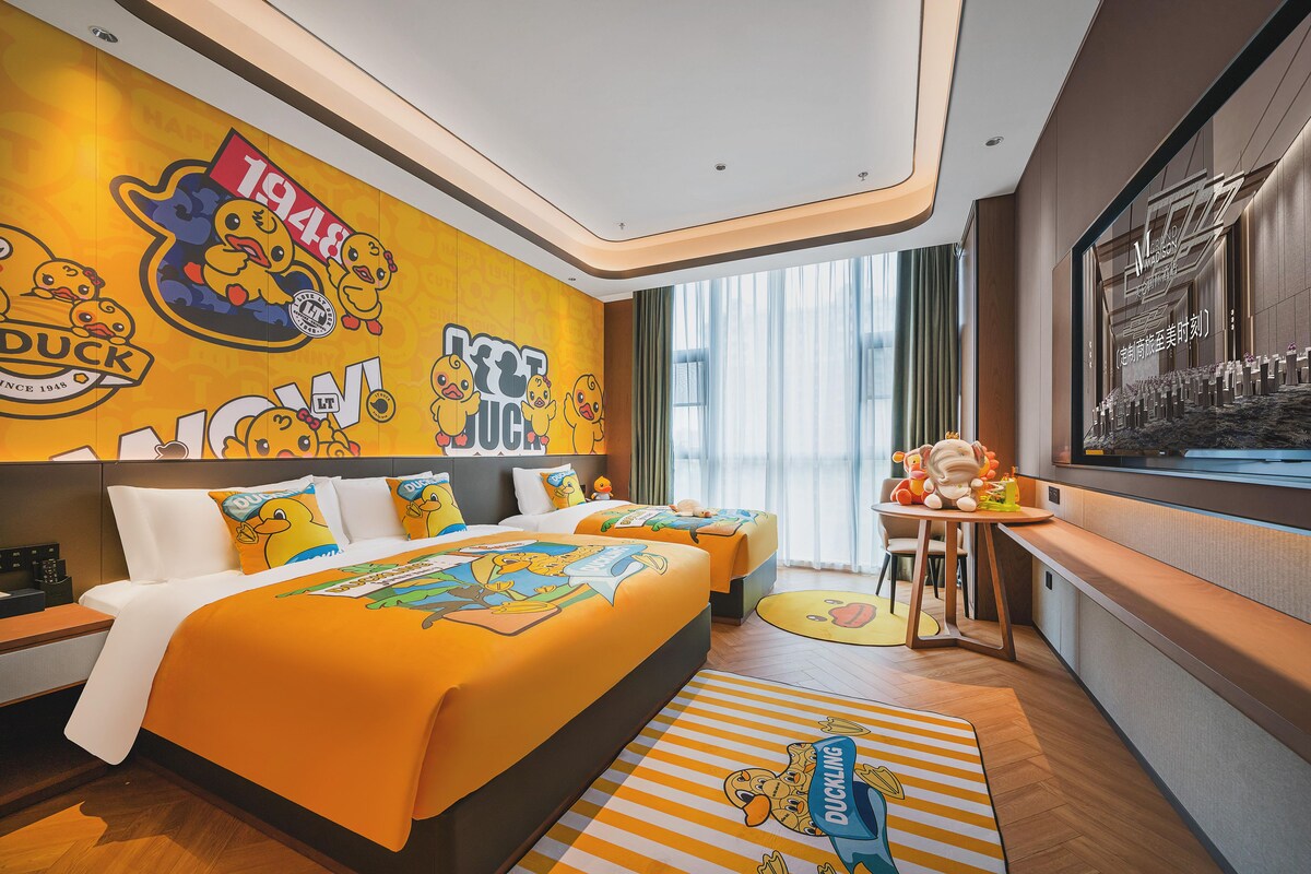 A hotel room with a duck-themed wall mural, a large bed with matching pillows, a desk with a chair, and a view of the city through large windows.