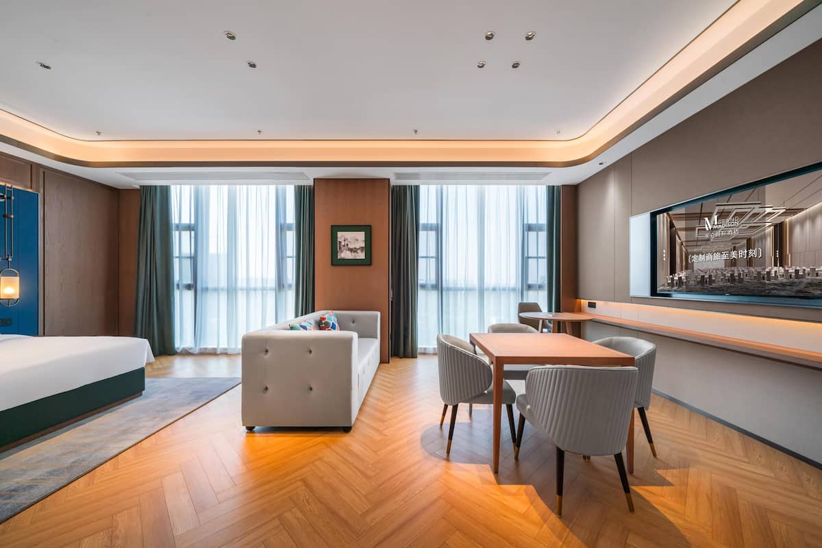 A modern hotel room with a bed, a sofa, a dining table with chairs, and a large window with curtains.