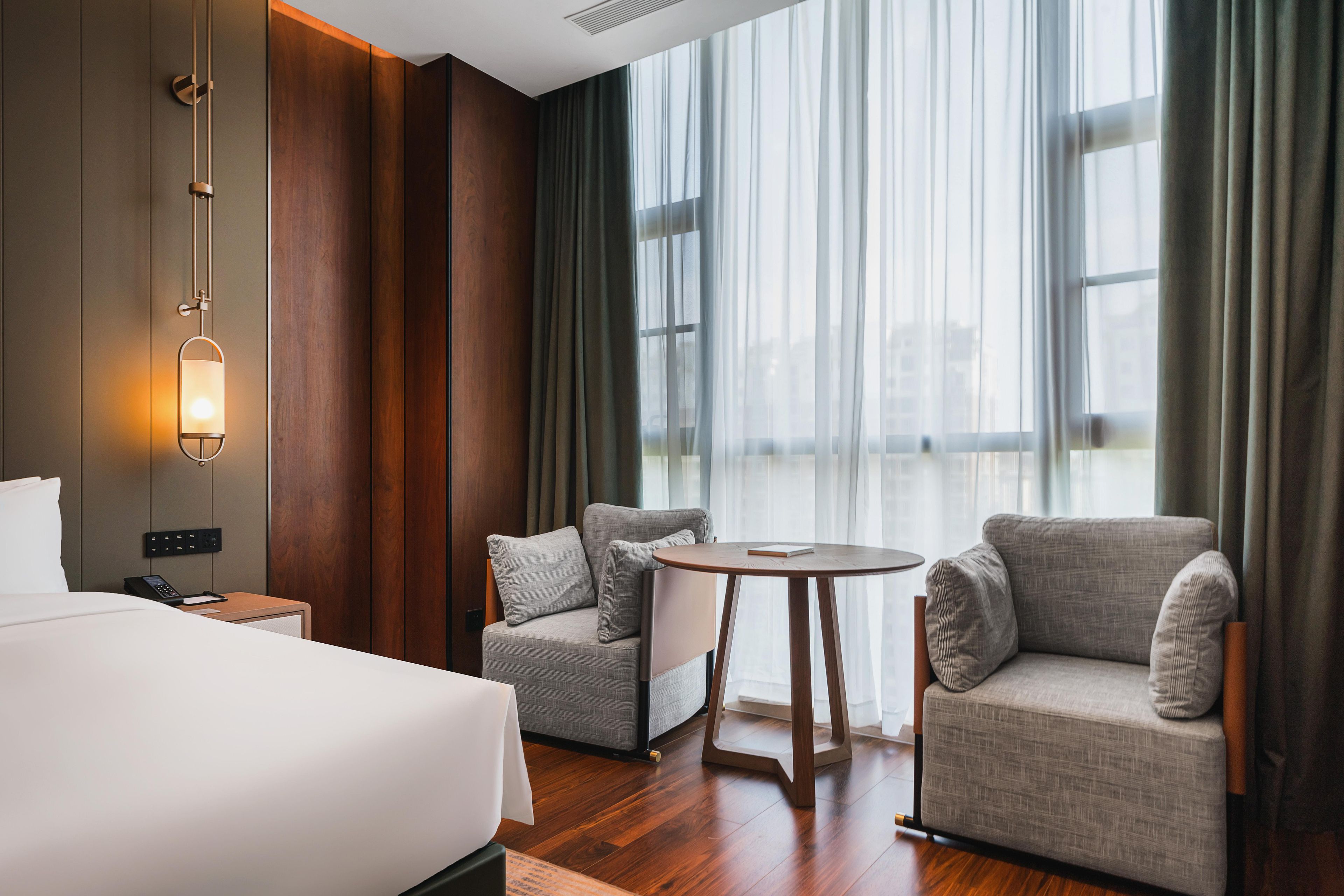 A hotel room with a bed, two armchairs, a small table, and a large window with curtains.