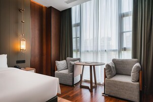Una habitación de hotel con una cama, dos sillones, una mesita y un ventanal grande con cortinas.