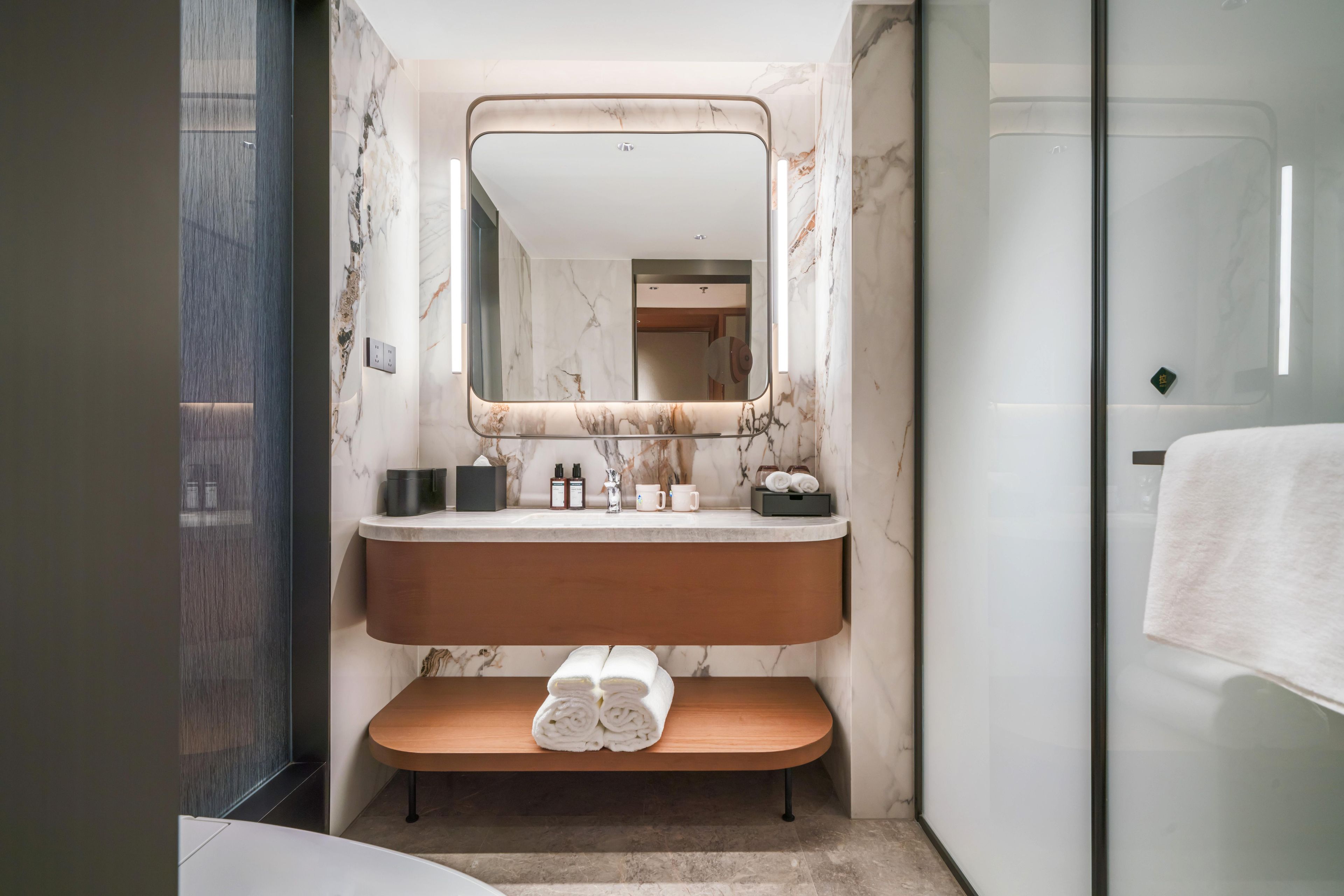 A modern bathroom with a marble vanity, a large mirror, and a bench with towels.