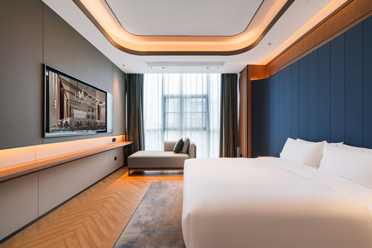 A modern hotel room with a large bed, a flat-screen TV mounted on the wall, a seating area with a sofa, and a large window providing a view of the city.