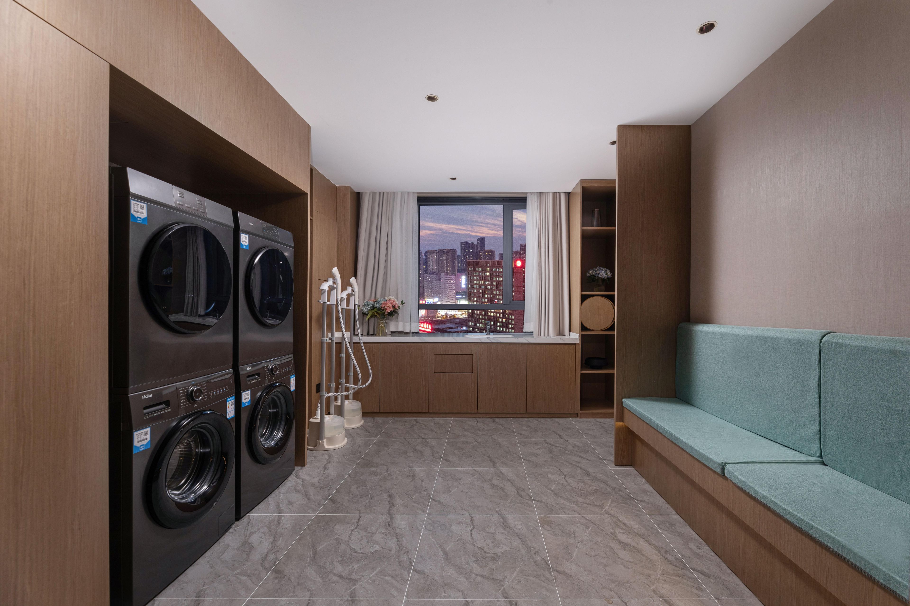 A modern laundry room with two washing machines, a window with curtains, a sofa, and a city view.