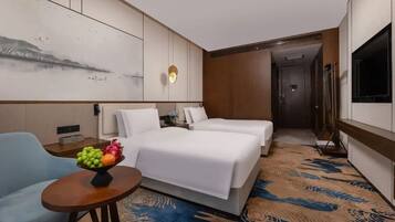 A hotel room with two beds, a small table with fruit, a painting on the wall, and a TV on the right.