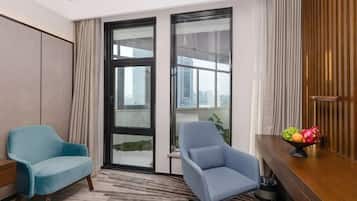 A hotel room with two blue armchairs, a wooden desk with a fruit bowl, and large windows offering a city view.