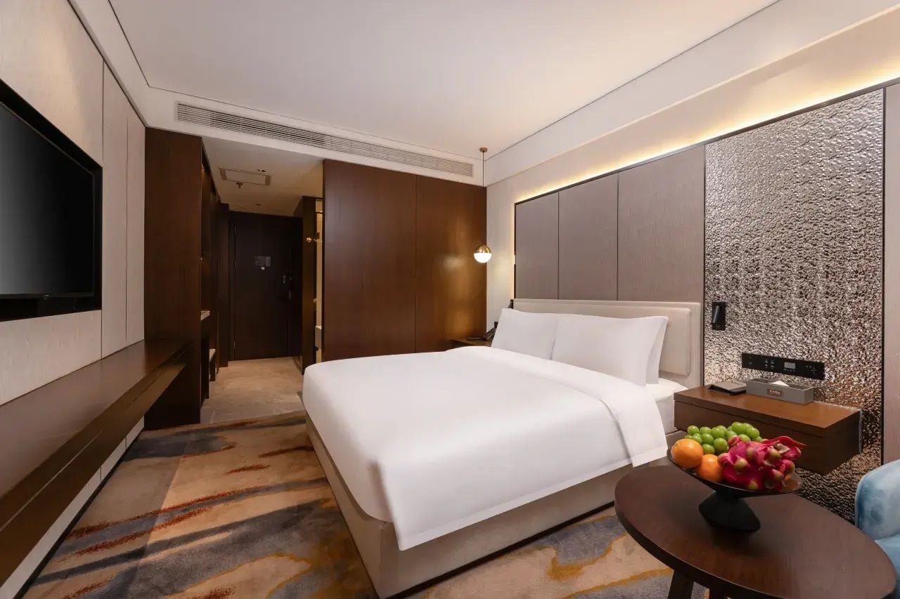 Una habitación de hotel con una cama grande, un televisor de pantalla plana, una mesita de noche con fruteros y un panel de control instalado en la pared.