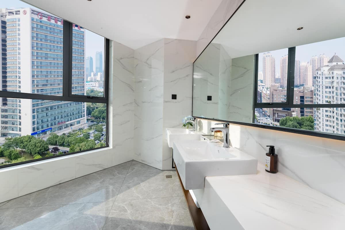 Un baño moderno con un espejo grande, dos lavamanos y vistas a un paisaje urbano a través de amplios ventanales.