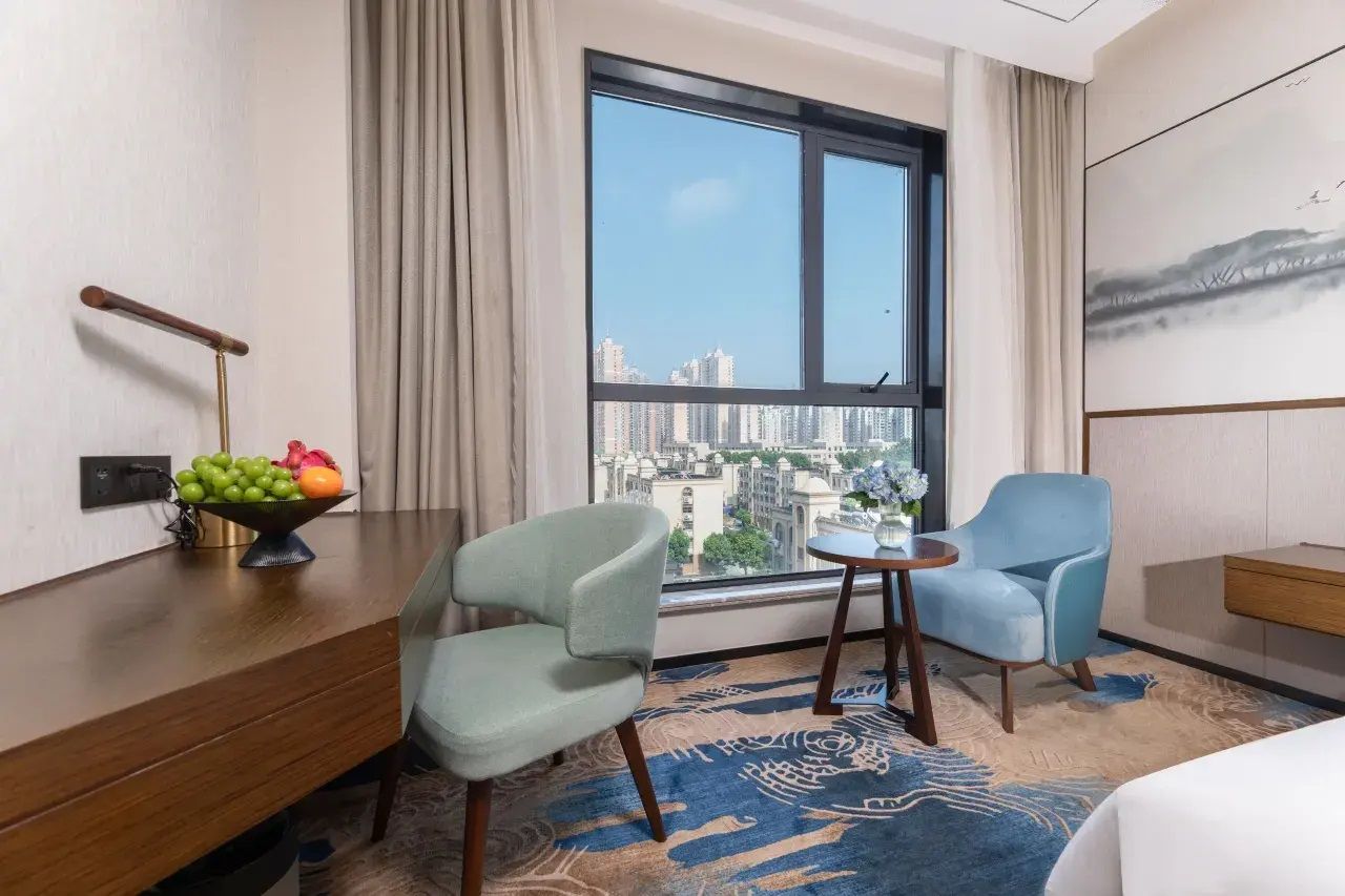 Una habitación de hotel con un ventanal grande, dos sillas, una mesita pequeña y un frutero sobre un escritorio de madera.