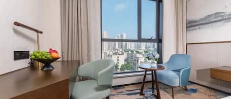 A hotel room with a large window, two chairs, a small table, and a fruit bowl on a wooden desk.