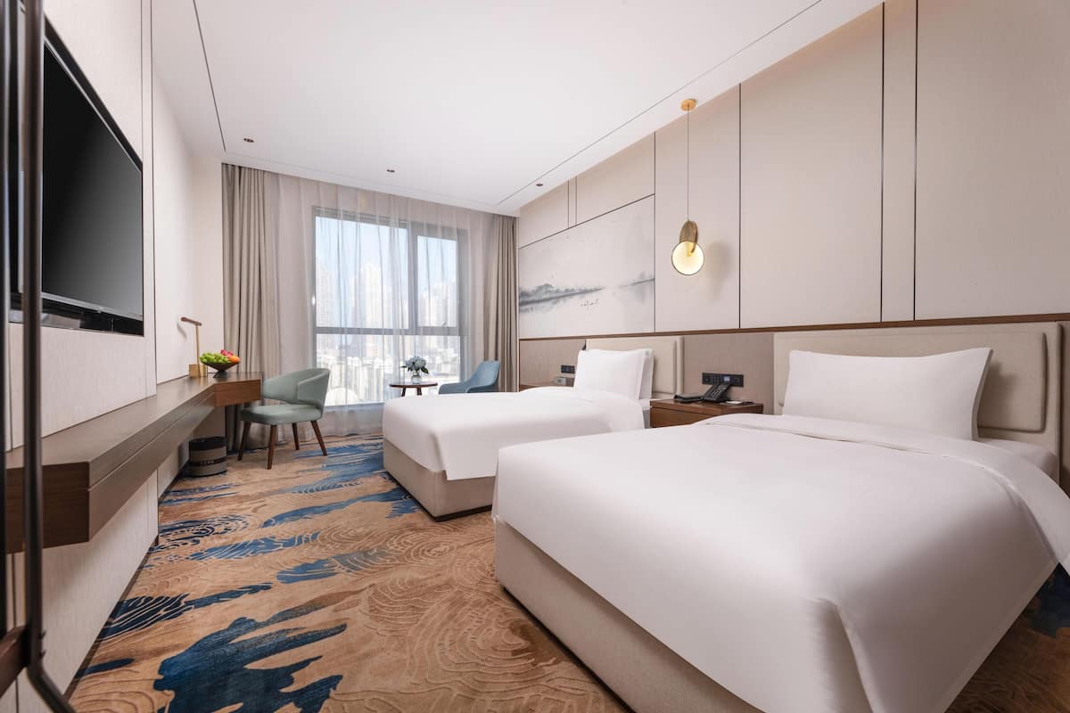 A hotel room with two beds, a desk, a chair, a TV, and a large window with curtains.
