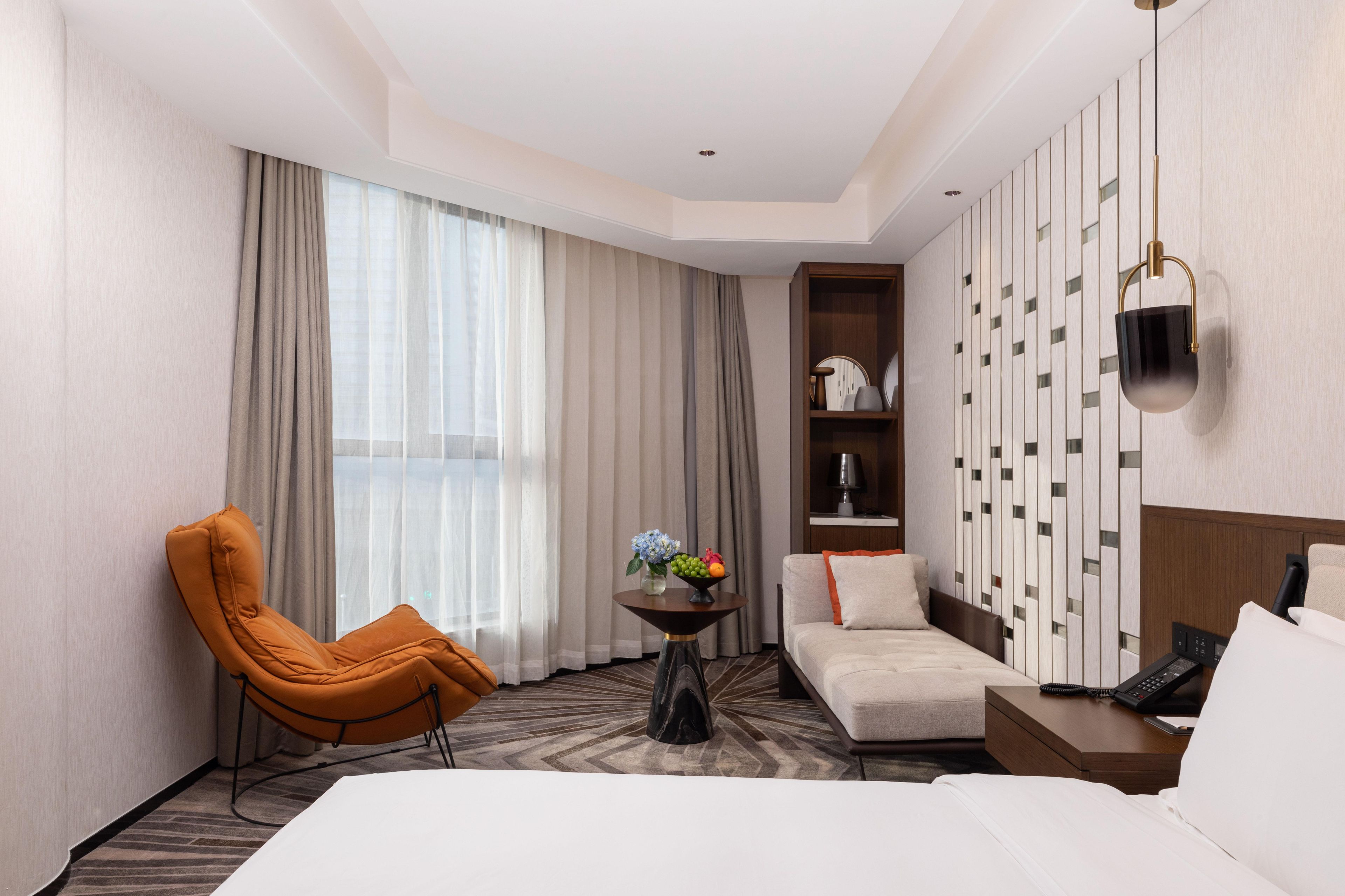 Una habitación de hotel con una cama, un sofá, una silla, una mesita con frutero, un teléfono en la pared y un panel decorativo.