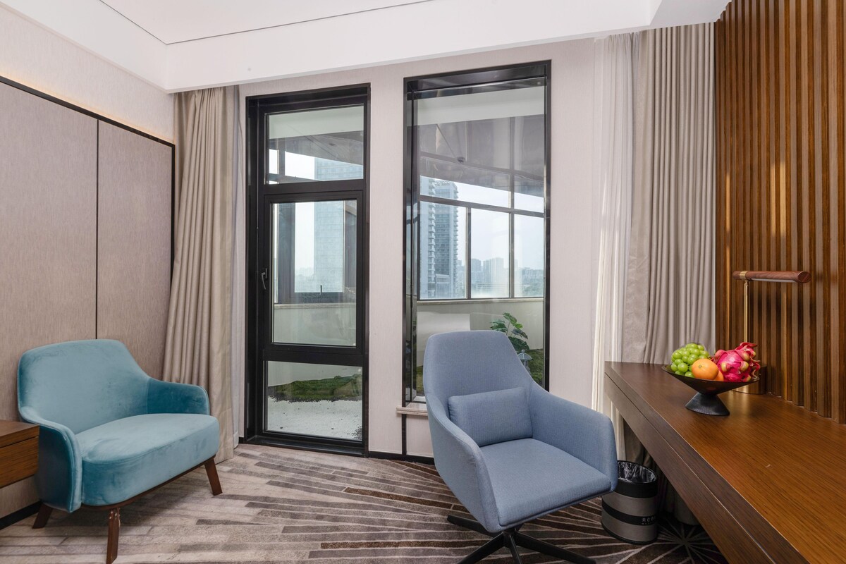 A hotel room with two blue armchairs, a wooden desk with a fruit bowl, a view of buildings through large windows, and a patterned carpet.
