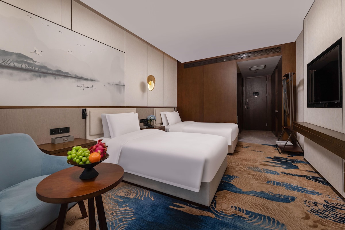 A hotel room with two beds, a small table with fruit, a TV, and a painting on the wall.