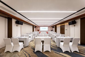 Un salón de conferencias con filas de mesas y sillas blancas, una pantalla grande al frente y altavoces en las paredes.