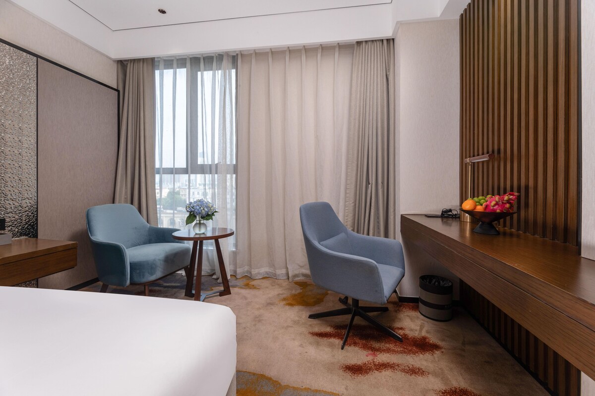 A hotel room with a bed, two armchairs, a small table with a vase, a fruit bowl, and a mirror on the wall.