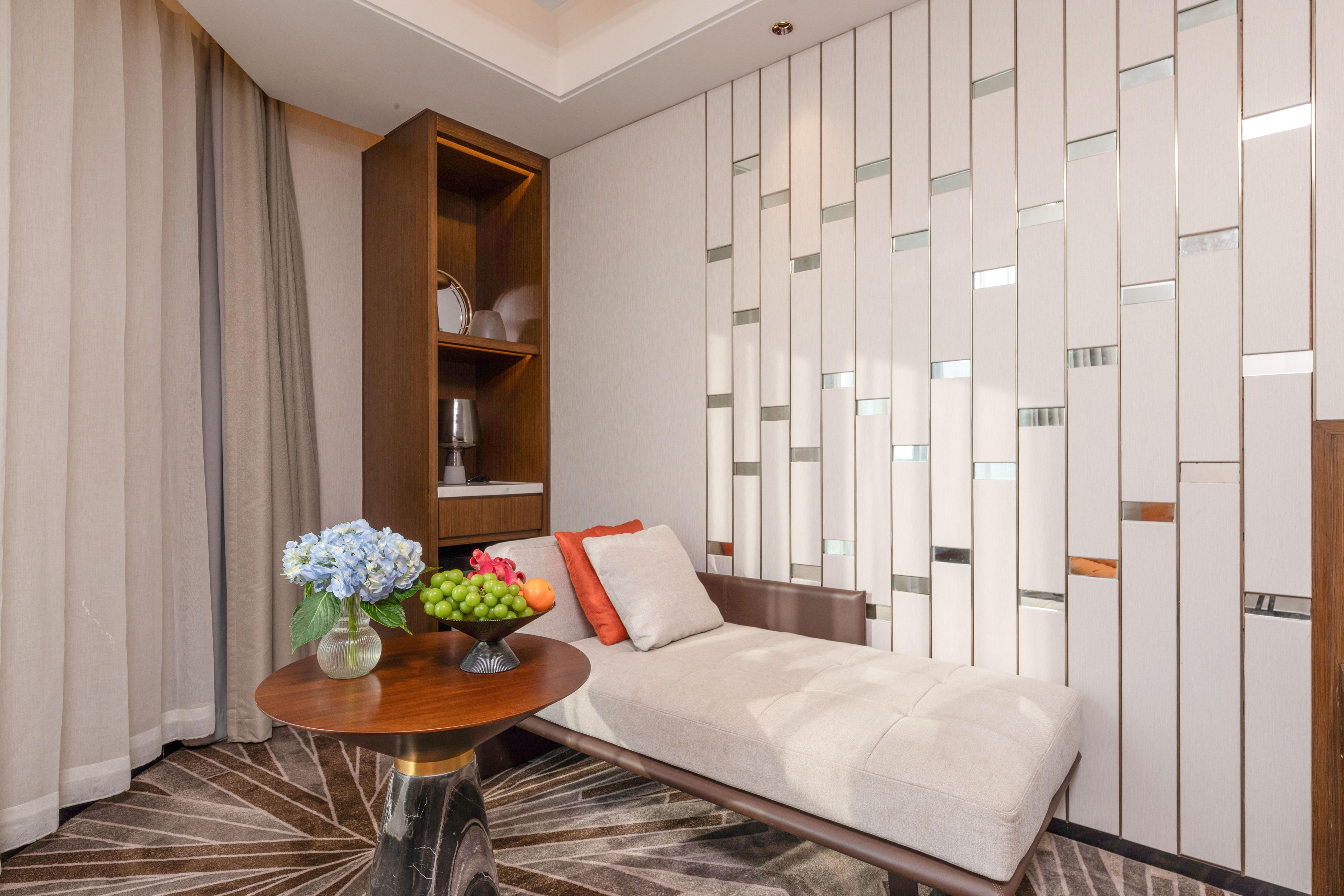 A modern hotel room with a sofa, a wooden side table with a vase of flowers, and a decorative wall panel.