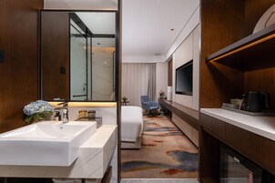 Una habitación de hotel moderna con un lavamanos grande de color blanco, una cama con cabecera blanca, un televisor montado en la pared y una mesita con un jarrón de flores.