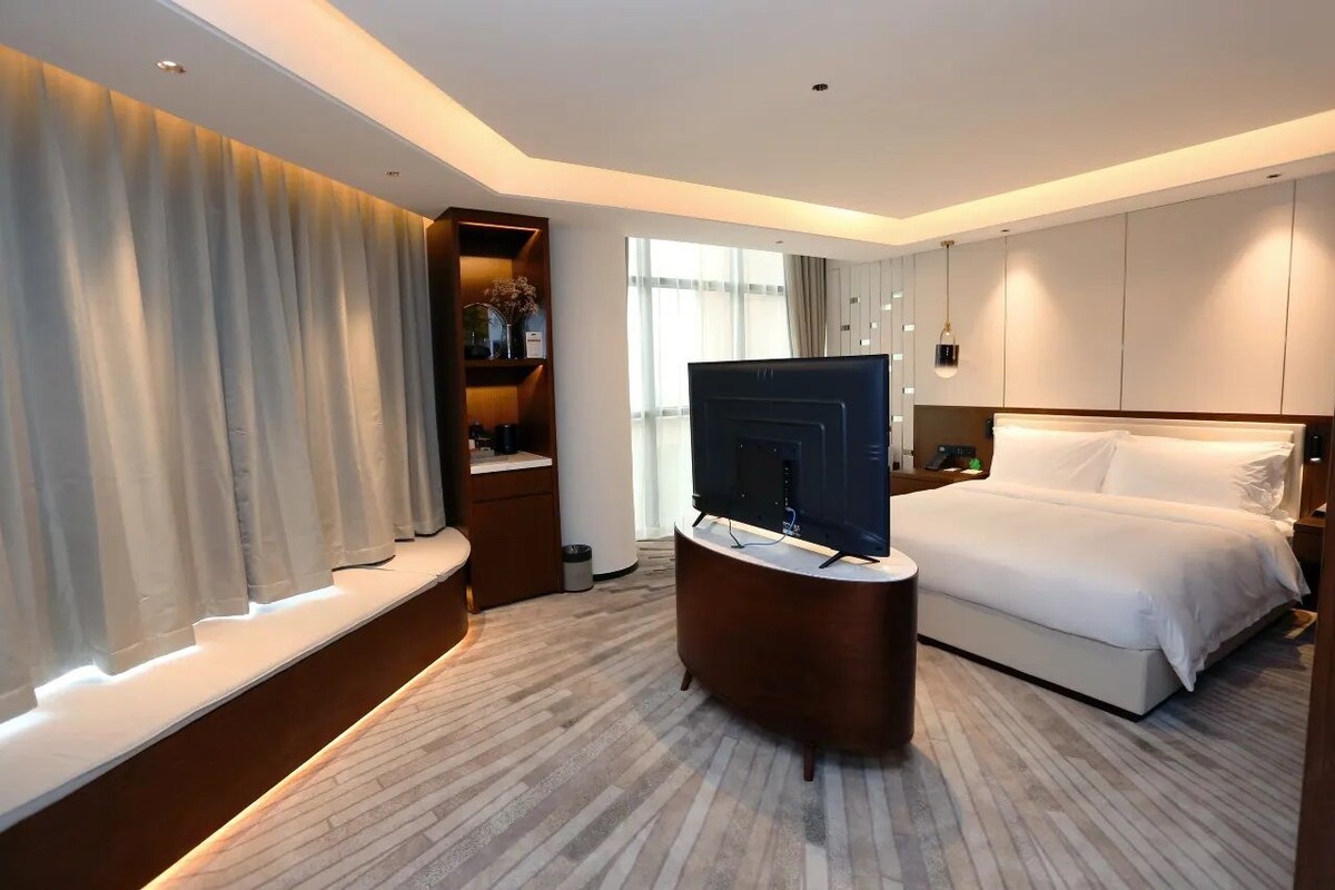 A hotel room with a bed, a TV on a stand, a bedside table, a wardrobe, and a window with curtains.