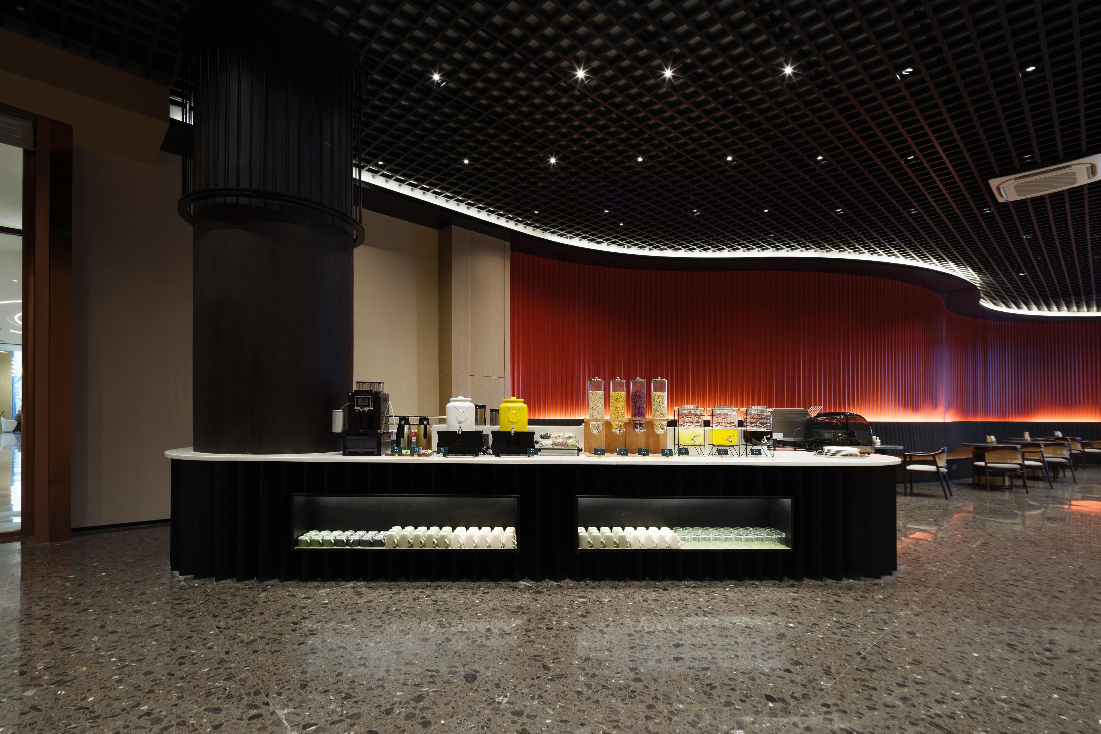 Un lobby moderno con una pared curva roja, una columna negra central y una zona de bar con diversos elementos.