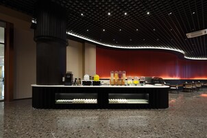 Un lobby moderno con una pared curva roja, una columna negra central y una zona de bar con diversos elementos.