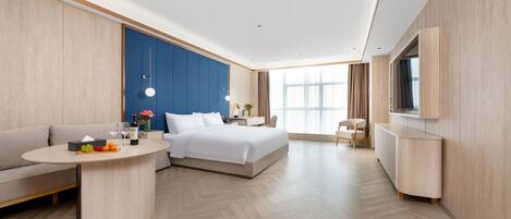 Una habitación de hotel moderna con una cama grande, un sofá, una pequeña mesa con fruta y una botella, un escritorio con una lámpara y un televisor en la pared.