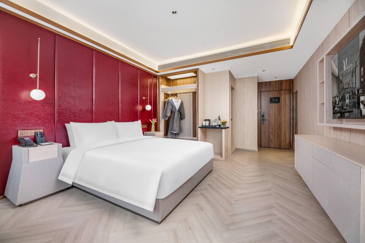 Una habitación de hotel moderna con una cama grande, una mesita de noche con teléfono, un armario, un tocador con espejo y una puerta que da a otra habitación.