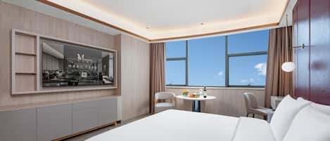 A modern hotel room with a large bed, a round dining table, a seating area, and a large window offering a view.