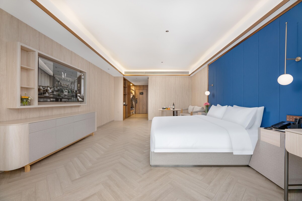 Una habitación de hotel moderna con una cama grande, un televisor montado en la pared, un escritorio con una silla y una pequeña mesa con lámpara.