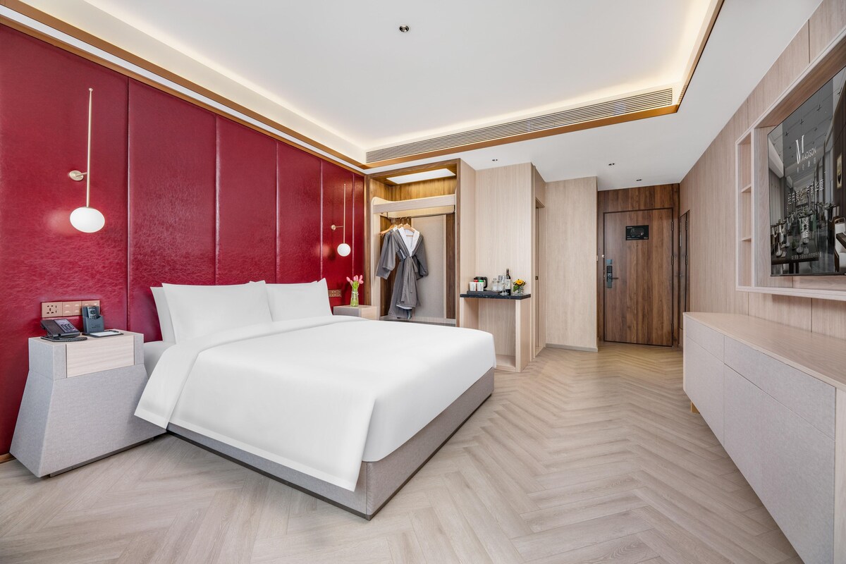 Ein modernes Hotelzimmer mit einem großen Bett, einem Nachttisch mit Telefon, einem Kleiderschrank, einem Schminktisch mit Spiegel und einer Tür zu einem weiteren Raum.